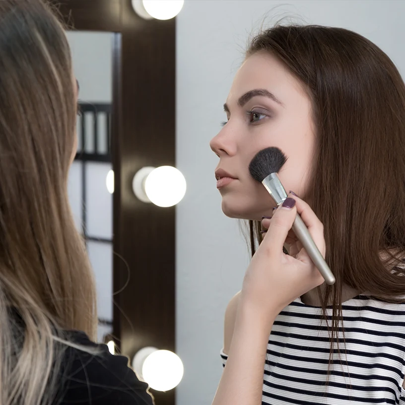 Une femme est maquillée au pinceau devant un miroir bordé de lumières.