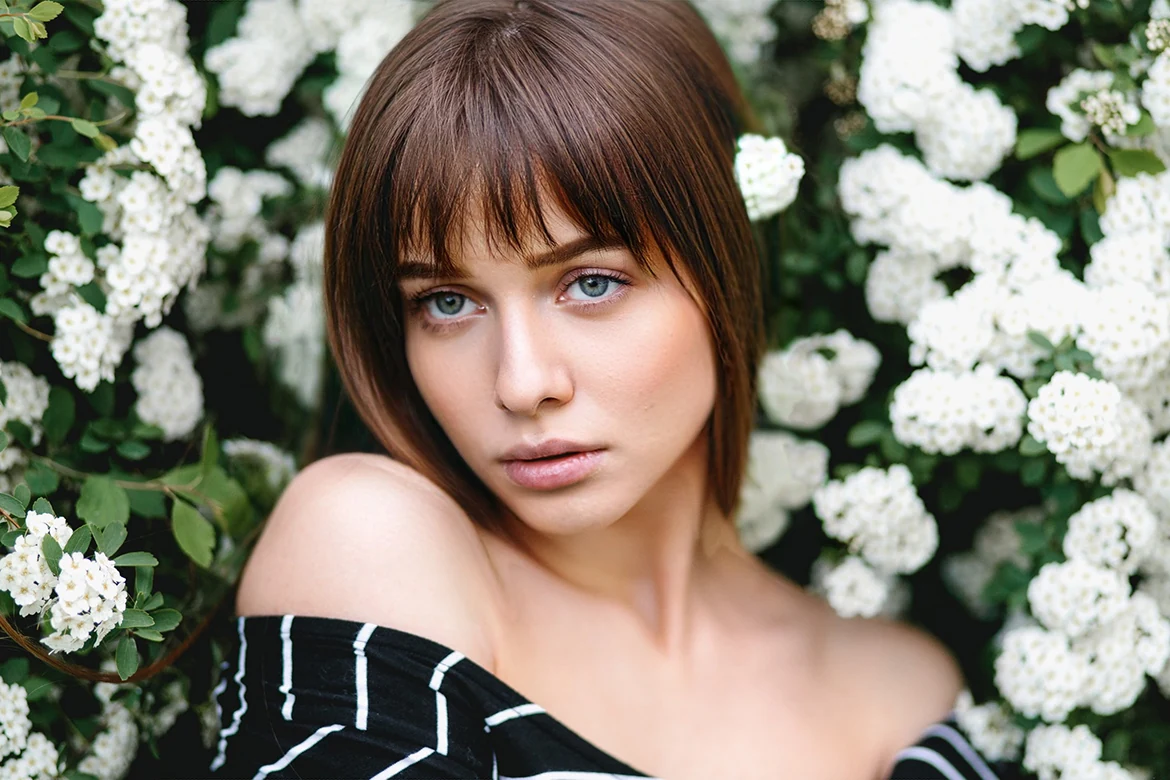 Portrait d’une femme brune à frange, devant des fleurs blanches.