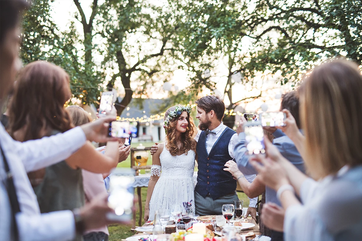 Un couple de mariés à table est entouré d’invités qui lèvent leur verre en plein air.