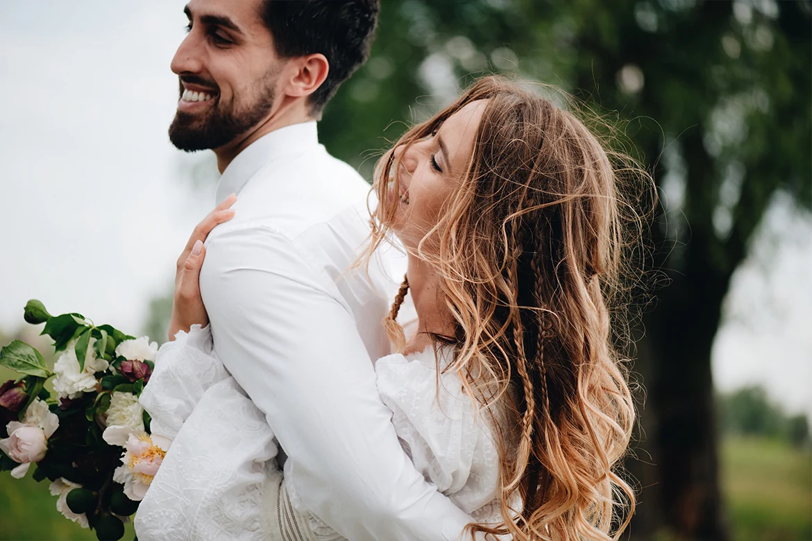Une femme en robe blanche enlace un homme en chemise blanche par-derrière, en extérieur.