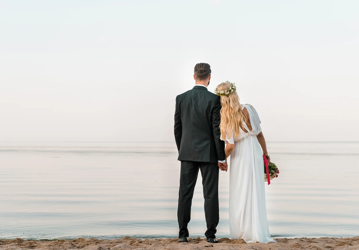 Un couple de mariés vu de dos se tient devant une mer calme, un bouquet à la main.