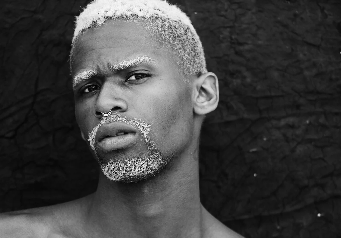 Portrait rapproché en noir et blanc d’un homme aux cheveux courts décolorés devant un mur sombre.