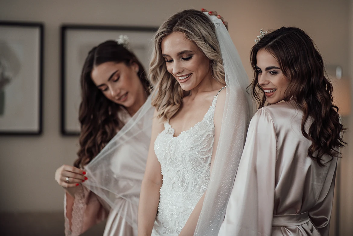 Une mariée en robe blanche se tient entre deux femmes en tenues claires pendant les préparatifs.