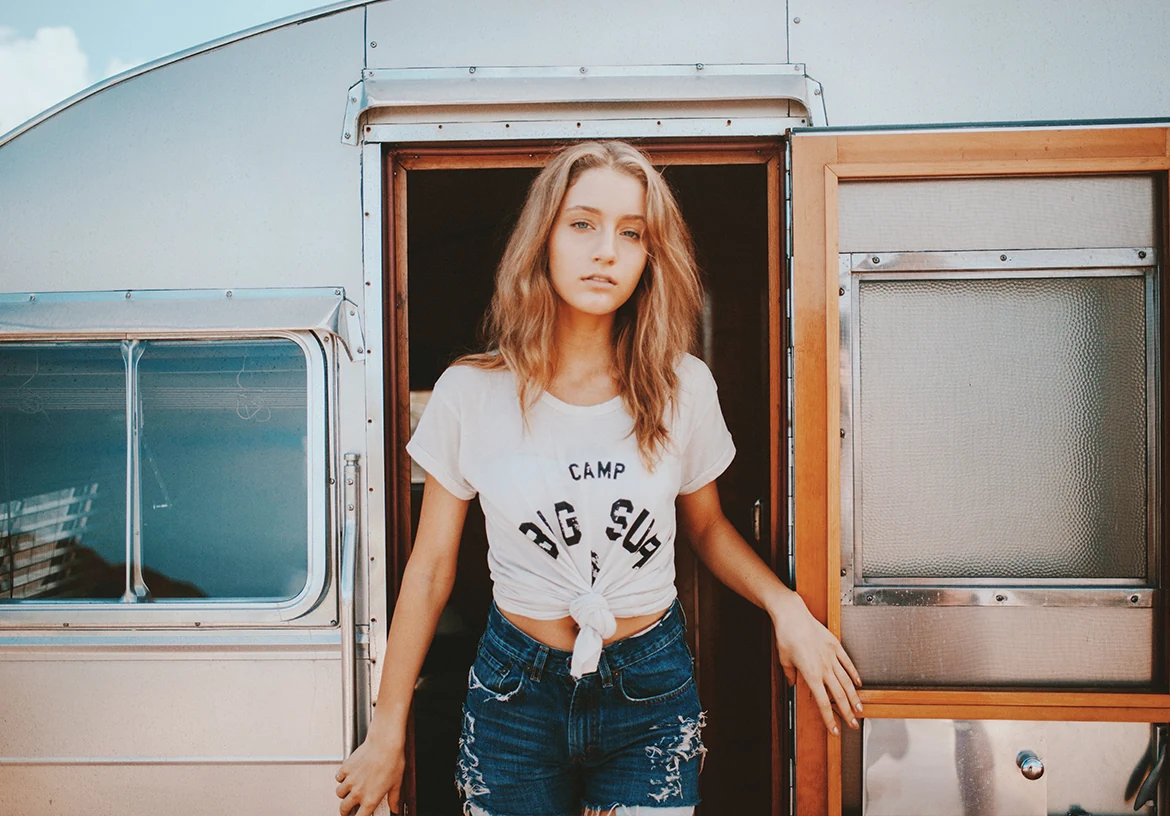 Une femme aux cheveux roux pose en tee-shirt blanc et jean devant une caravane.