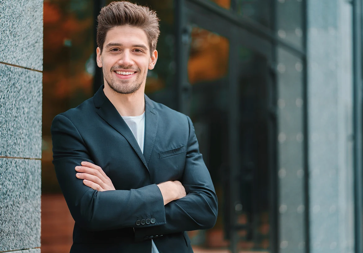 Un homme souriant en veste bleue pose les bras croisés devant une façade vitrée.