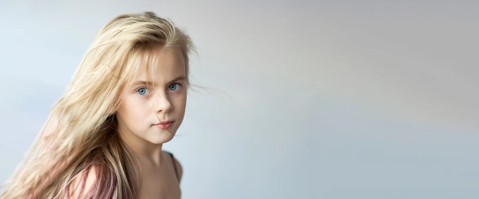 Portrait d’une jeune fille aux cheveux blonds sur fond gris clair, avec un large espace libre à droite.