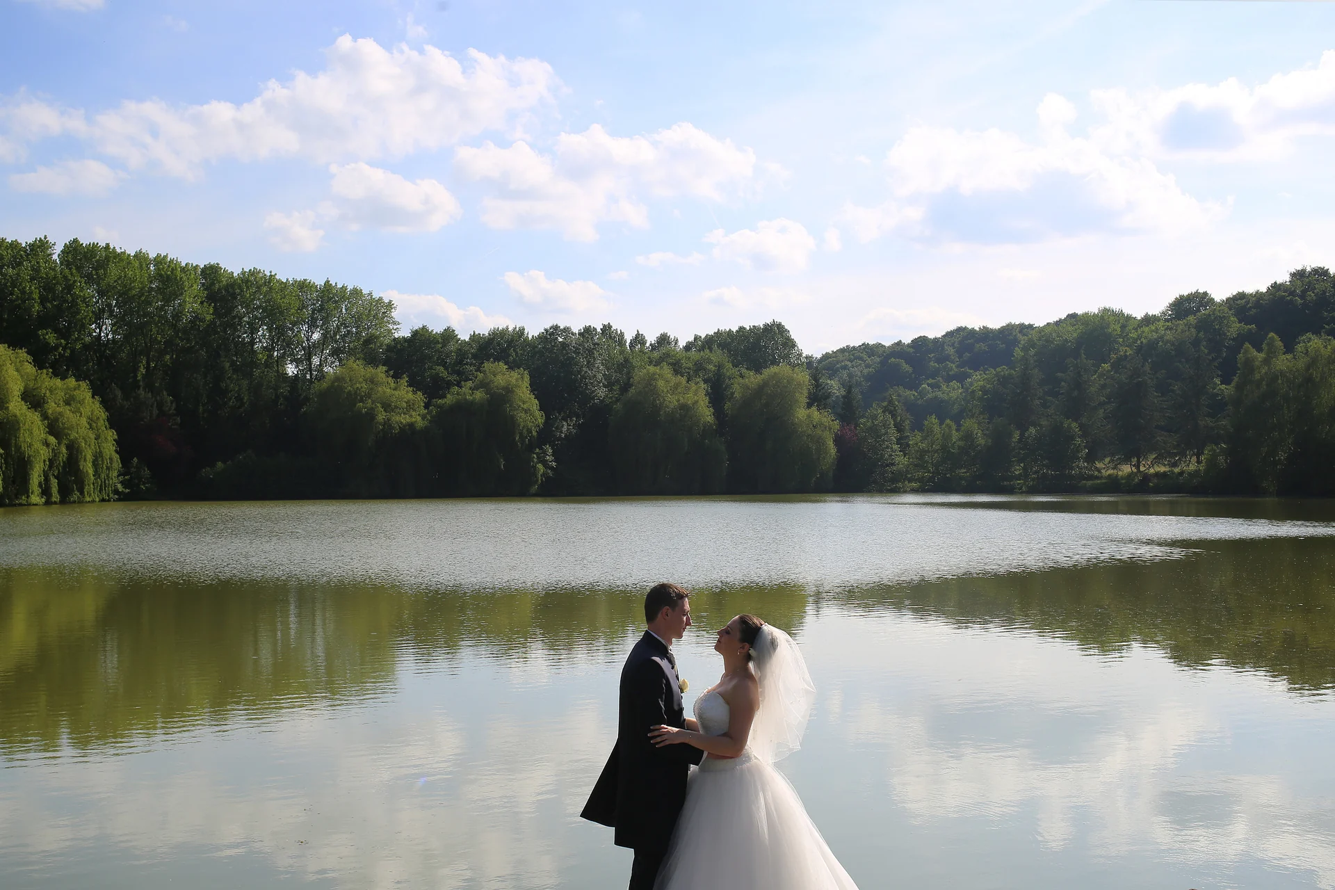 Les mariés face à face au bord d’un lac calme, avec le reflet des arbres dans l’eau