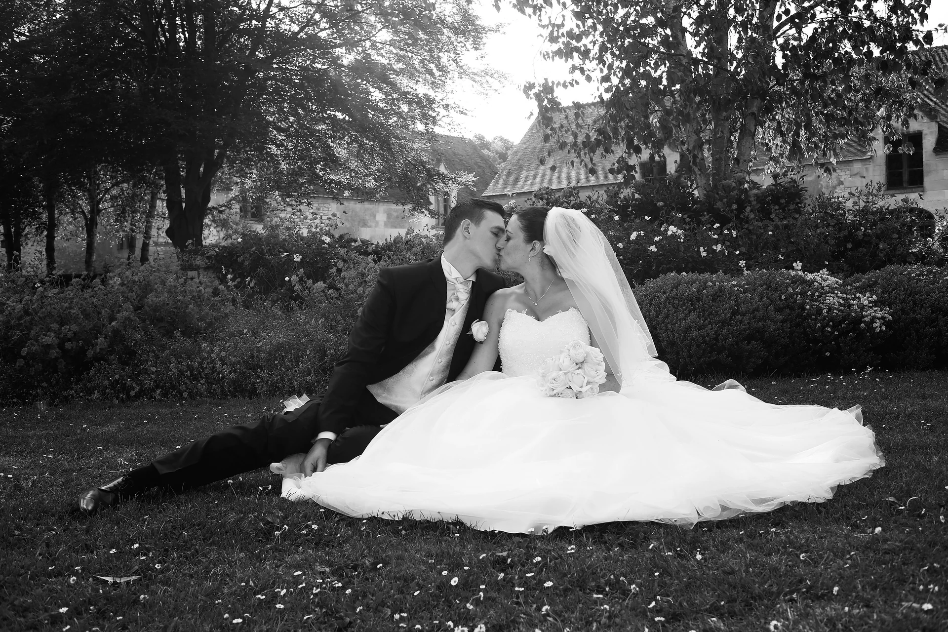 Les mariés s’embrassent assis dans un jardin, la robe déployée sur l’herbe, en noir et blanc