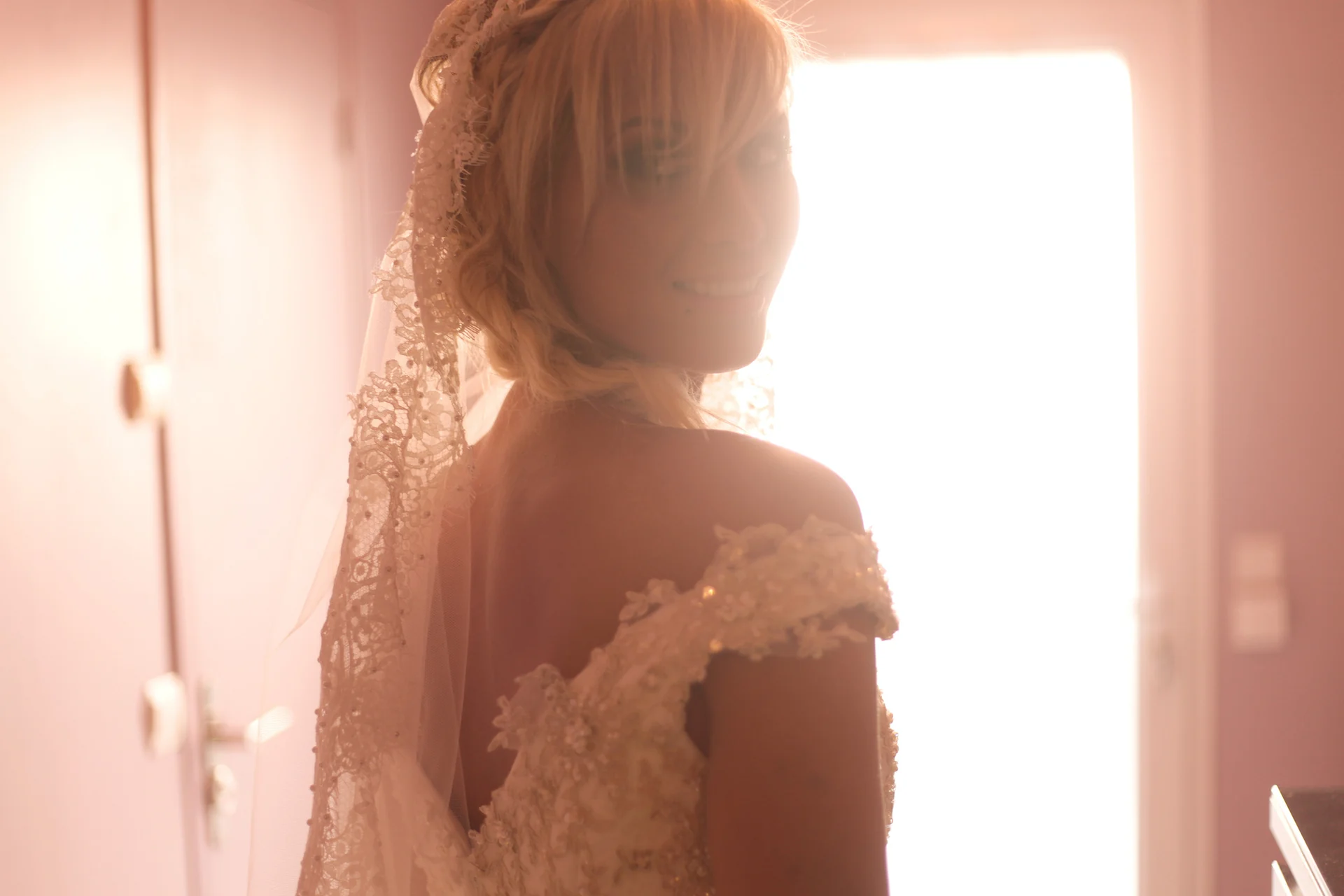 Une mariée en robe de dentelle et voile, éclairée à contre-jour devant une porte